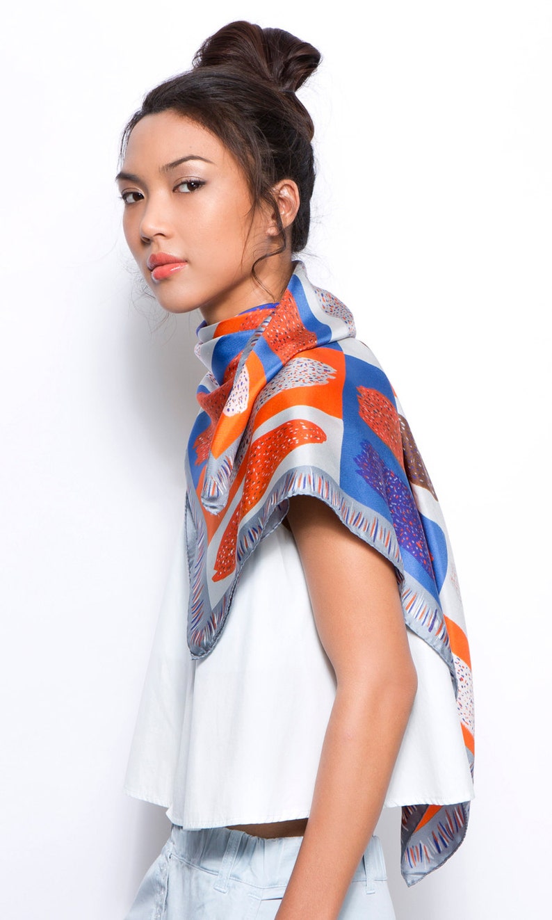 May include: A woman wearing a white top and blue jeans with a blue, orange and white patterned scarf tied around her neck.