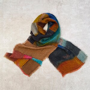 May include: A colorful patchwork scarf with a variety of hues including brown, blue, orange, and yellow. The scarf has a textured appearance and is draped on a neutral background. The scarf is a fashion accessory.