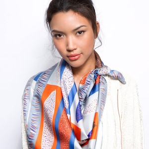 May include: A woman wearing a white cardigan with a blue, orange and grey patterned scarf. The scarf has a geometric design with abstract shapes.