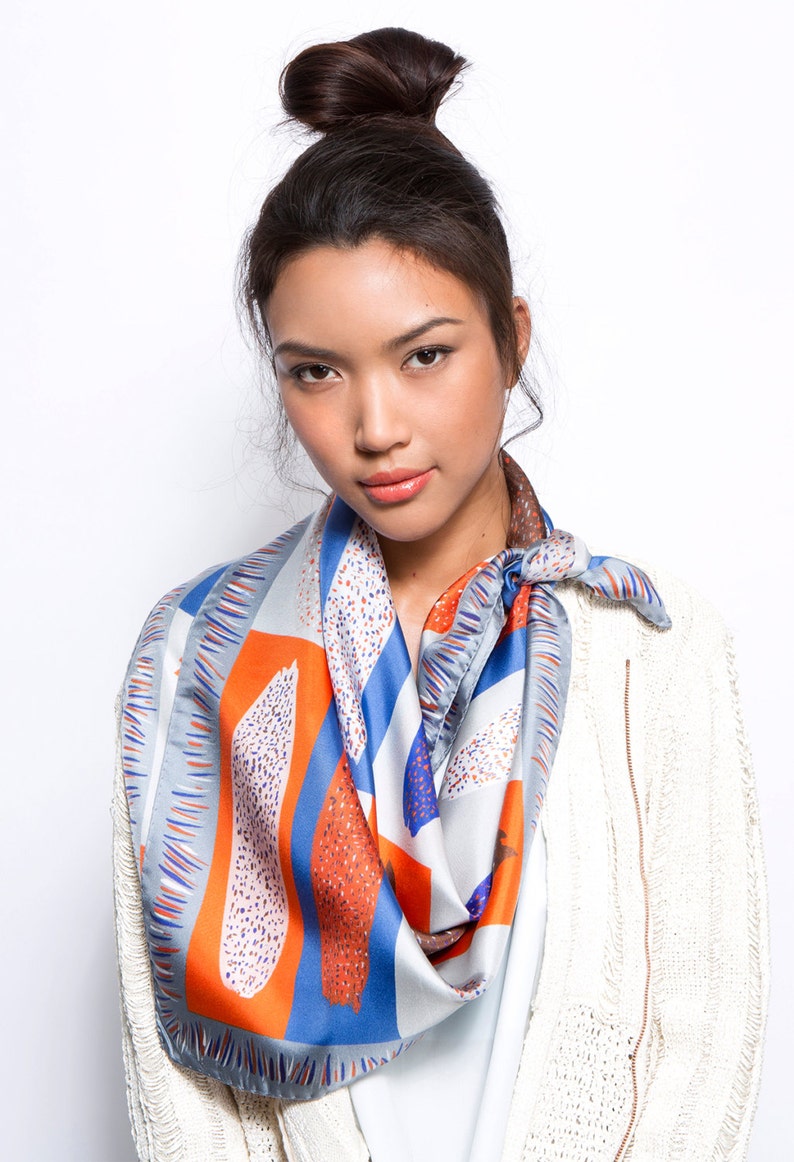 May include: A woman wearing a white cardigan with a blue, orange and grey patterned scarf. The scarf has a geometric design with abstract shapes.
