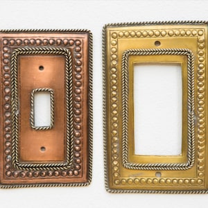 Puede incluir: Dos cubiertas decorativas de metal para interruptores de luz. Una es de color cobre con una abertura para un solo interruptor, y la otra es dorada con una abertura más grande. Ambas tienen un borde con cuentas y un ribete trenzado similar a una cuerda.