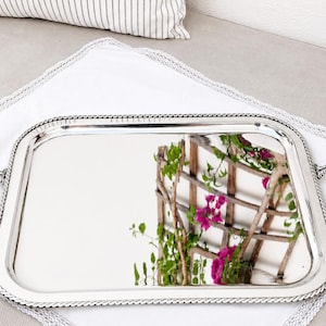 Può includere: Un vassoio da portata rettangolare color argento con una superficie lucida e riflettente. Il vassoio presenta un bordo decorativo a corda e due manici decorati e strutturati sui lati opposti, dal design classico.