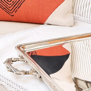 Puede incluir: Una bandeja de servicio rectangular de plata con una superficie pulida y reflectante y asas decorativas ornamentadas. La bandeja presenta un detalle de borde con cuentas a lo largo del perímetro, mostrando un acabado metálico que refleja la tela naranja y negra circundante.