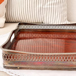 Könnte beinhalten: Ein rechteckiges Serviertablett mit einer polierten, dunkel rötlich-braunen Holzmaserung als Boden und einer silberfarbenen Metallgalerie. Der Metallrand zeigt ein komplexes Gittermuster an den Seiten sowie integrierte Griffe.