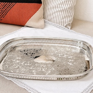 May include: An oval silver-coloured serving tray featuring an intricate, embossed floral and scrollwork pattern across its base. The tray is designed with a raised, openwork gallery rail along the entire perimeter, providing a decorative border.