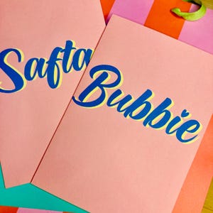 May include: Two pink cards with the words "Saft" and "Bubbie" written in blue and yellow lettering.