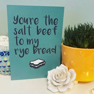 May include: A teal card with the text "You're the salt beef to my rye bread" in black lettering. A simple illustration of a sandwich is below the text. The card is sitting on a table with a white rose and a yellow pot with a green succulent plant.