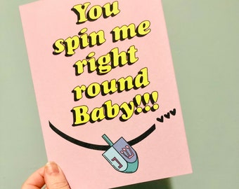May include: Pink card with the text "You spin me right round Baby!!!" in yellow letters with a black string and a blue and purple dreidel with a white "1" on it.