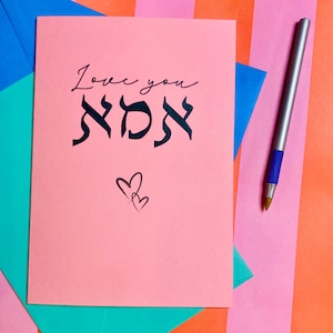 Könnte beinhalten: Eine rosa Karte mit den Worten "Love you" in Kursivschrift und dem hebräischen Wort für "Mutter" in Kalligraphie. Zwei kleine Herzen sind in der rechten unteren Ecke gezeichnet.