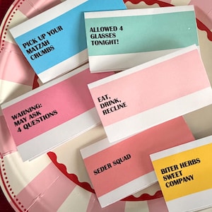 May include: A collection of colorful cards with various phrases printed on them, including "Pick Up Your Matzah Crumbs" and "Allowed 4 Glasses Tonight!" The cards are in shades of blue, pink, and yellow, with white borders.