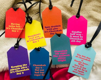 May include: Eight colorful gift tags with text about the eight days of Hanukkah. The tags are tied with black ribbon and are arranged on a fuzzy white surface.