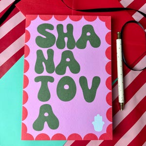 May include: A rectangular greeting card with a light pink background and a scalloped red border. The card features the dark green text SHANA TOVA in a bold, rounded font, accompanied by a small white hamsa symbol at the bottom right corner.