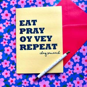May include: A yellow greeting card with the text "EAT PRAY OY VEY REPEAT" in navy blue, with "chag sameach" written below. A white pen with silver accents and a polka dot pattern rests on the card. The background features a floral pattern.