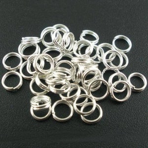May include: A pile of small, silver split rings, spread out on a dark background. These metal rings are designed to be opened and closed, often used in jewellery making, keychains, and craft projects. The rings are circular and have a split in the metal.