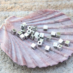 May include: Small, silver cube beads, each with a central hole, are scattered on a large, pink seashell. The beads are set against a sandy background. These beads, likely for jewellery making, are approximately 0.5 cm in size.