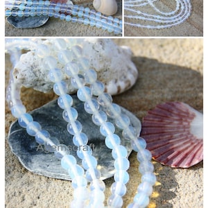 May include: Several strands of opalescent, round gemstone beads are shown on a sandy surface with seashells. The beads have a soft, milky white colour with a subtle iridescence. The image includes close-up shots of the beads.
