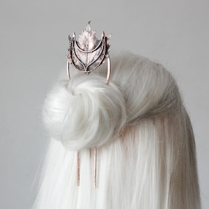 Op de afbeelding: Een koperen haar kam met een bladontwerp en een halve maan vorm. De kam wordt gebruikt om een ​​knot van wit haar vast te houden.