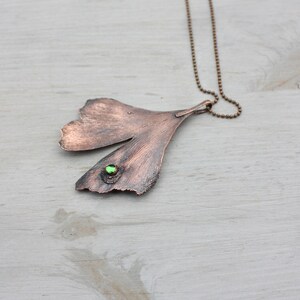 May include: A copper ginkgo leaf pendant necklace with a green gemstone accent. The necklace is on a silver chain.