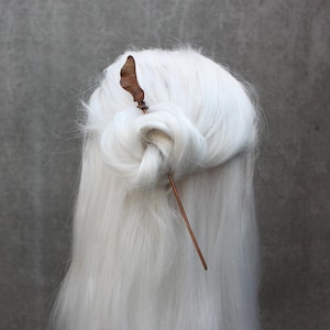 May include: A copper-colored hair stick with a leaf-shaped top is inserted into a bun of long, white hair. The hair is styled in a loose, elegant updo, with the stick adding a natural, earthy accent. The background is a solid, neutral gray.