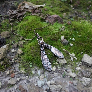 May include: A handcrafted necklace featuring a pendant shaped like two silver maple seeds. The pendant hangs from a delicate chain, resting on a bed of green moss and small rocks. The seeds have a textured, metallic finish.