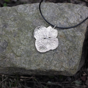 May include: A silver pendant necklace with a three-lobed flower design. The pendant is suspended from a black cord. The necklace is displayed on a textured grey stone, with a blurred natural background.