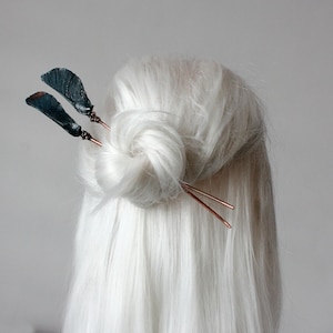 Op de afbeelding: Twee donkerblauwe veren haarspelden met koperen draadaccenten. De spelden worden gebruikt om een ​​knot van wit haar vast te houden.