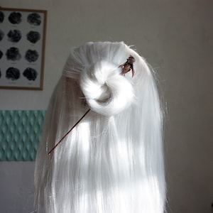 May include: White hair styled in a bun, secured with a brown hair stick and a decorative element. The long, straight hair has a natural, flowing look. The bun is centrally positioned, with the hair stick inserted through it. The brown decorative element adds a unique touch to the hairstyle.