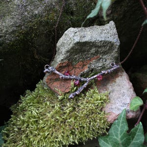 Op de afbeelding: Een zilveren ketting met een takontwerp en rode edelsteenaccenten. De ketting wordt getoond op een mossige rots.