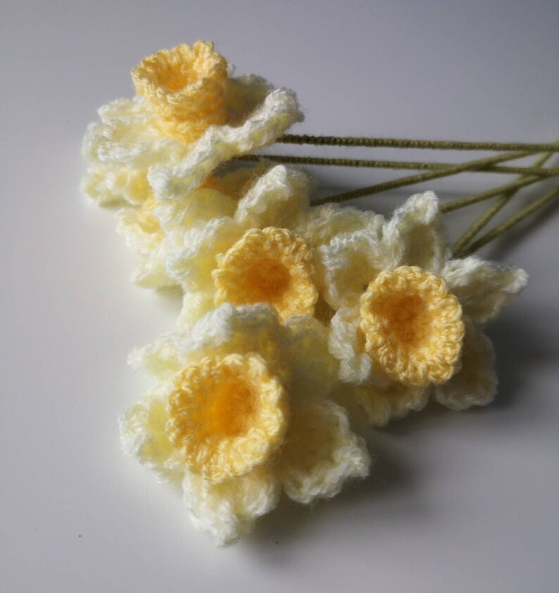 May include: A bunch of crocheted daffodils in shades of yellow and white. The flowers are arranged in a bouquet and have green stems.