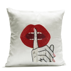 Può includere: Cuscino bianco con un design di labbra rosse perlate e un dito nero sulle labbra, a simboleggiare il silenzio.