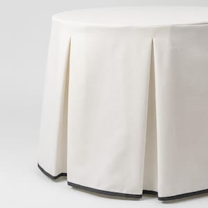 May include: A round, off-white fabric table cover featuring a pleated skirt design that extends to the floor. The bottom edge of the fabric is finished with a thin, dark charcoal gray trim that outlines the base of the pleated panels.