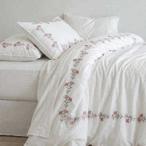 Op de afbeelding: Een wit beddengoedset bestaande uit een dekbedovertrek en twee kussenslopen, allemaal versierd met een geborduurd patroon van kleine roze bloemen en groene stengels in een rechthoekige rand op de stof.