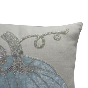 May include: A decorative pillow with a light gray background and a blue pumpkin design. The pumpkin is embellished with silver beads, and the stem and vine are also beaded. The pillow has a soft, textured appearance.