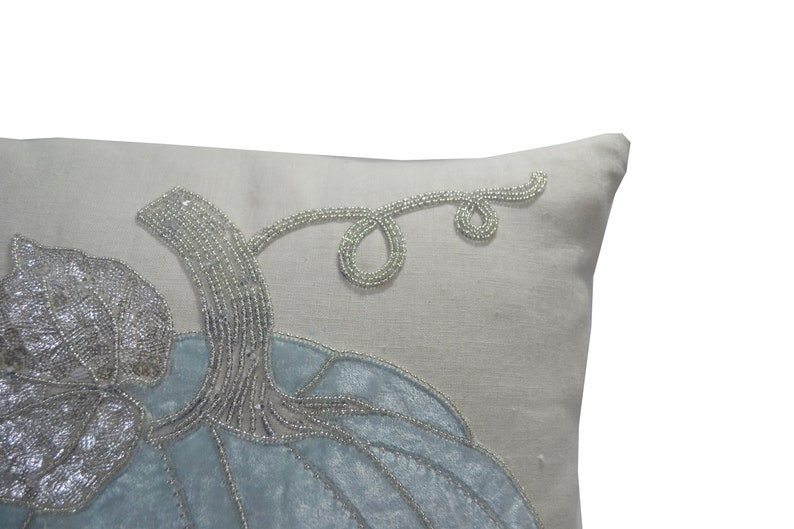 May include: A decorative pillow with a light gray background and a blue pumpkin design. The pumpkin is embellished with silver beads, and the stem and vine are also beaded. The pillow has a soft, textured appearance.