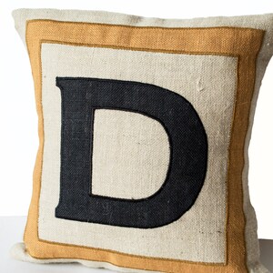 May include: A beige pillow with a thick, orange border and a black embroidered letter "D" in the center.