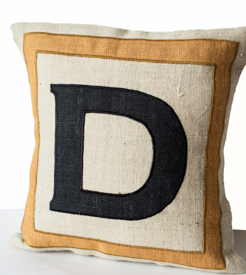 May include: A beige pillow with a thick, orange border and a black embroidered letter "D" in the center.
