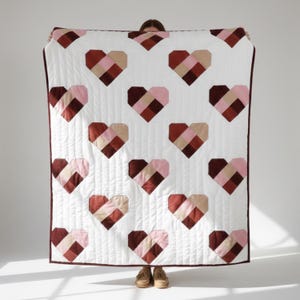 May include: A white quilt featuring a repeating pattern of geometric hearts in shades of pink, beige, and burgundy. The quilt has a burgundy border and is held up against a white background.