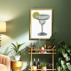 May include: A gold bar cart with a glass top and two shelves. The cart is decorated with a brown leather strap. The cart is filled with bottles of liquor, glasses, and plants. A framed print of a margarita cocktail hangs on the wall behind the cart.