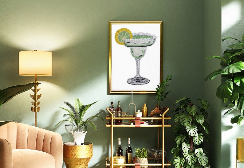 May include: A gold bar cart with a glass top and two shelves. The cart is decorated with a brown leather strap. The cart is filled with bottles of liquor, glasses, and plants. A framed print of a margarita cocktail hangs on the wall behind the cart.