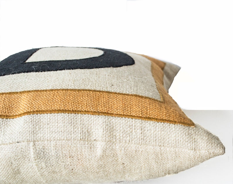 May include: A white linen pillow with a black and mustard yellow geometric design. The pillow is shown from the side, with the seam visible.