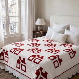 May include: A white quilt with a repeating pattern of red house shapes. The quilt is on a bed with white pillows and a white bed skirt. The room has a window with white curtains and a small wooden side table.
