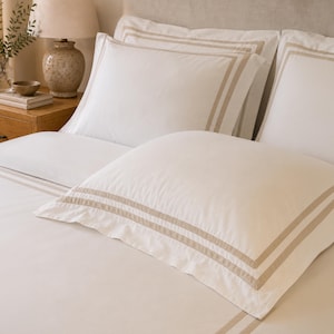 Puede incluir: Un juego de ropa de cama blanco que incluye dos almohadas cuadradas grandes y una sábana, cada una con rayas dobles de color beige en los bordes. La tela lisa cubre la cama, creando una apariencia limpia y uniforme con fundas a juego.