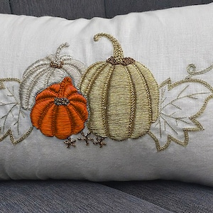 May include: A rectangular white pillow with an embroidered design of three pumpkins and leaves. The pumpkins are in shades of orange, silver, and gold, with gold beaded accents. The pillow is placed on a gray surface.