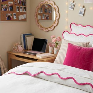 May include: A white bedding set featuring a scalloped edge with bright pink trim. The set includes a duvet cover and pillow shams, all accented with the same pink scalloped detailing. The bedding is displayed on a bed with a solid pink throw pillow.