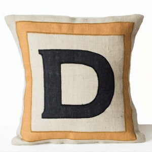 May include: A beige pillow with a large black embroidered letter "D" in a double-lined orange frame.