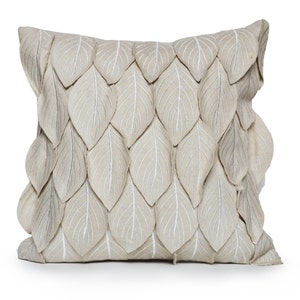 Puede incluir: Un cojín decorativo cuadrado con una funda de tela beige. El cojín está adornado con numerosas hojas de tela beige, cada una con detalles de venas blancas, creando un efecto texturizado y en capas. Diseñado para la decoración del hogar.