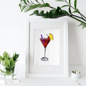 May include: A framed beaded artwork of a red cocktail with a cherry and pineapple garnish. The cocktail is in a martini glass with a purple straw.