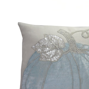 May include: A decorative throw pillow with a light blue pumpkin design. The pumpkin is embellished with silver sequins and beads, and features a stem and leaf. The pillow has a light gray background.