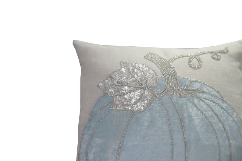May include: A decorative throw pillow with a light blue pumpkin design. The pumpkin is embellished with silver sequins and beads, and features a stem and leaf. The pillow has a light gray background.