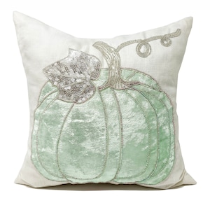 May include: A cream-coloured cushion featuring a light green velvet pumpkin design. The pumpkin is decorated with silver sequins and beads. The cushion is a seasonal accent for autumn, and is suitable for a sofa or bed.